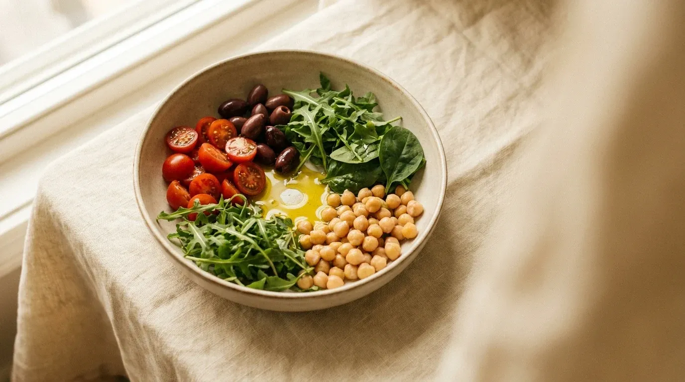 Overhead view of a ceramic bowl filled with Mediterranean salad of tomatoes, olives, greens, chickpeas, and olive oil.